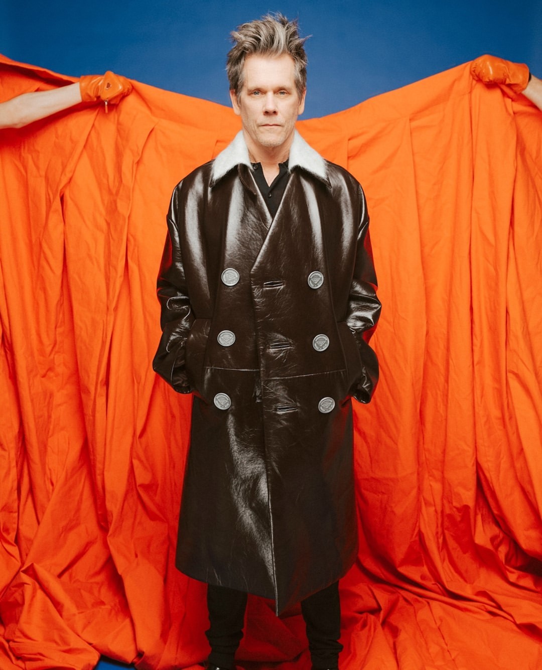 Portrait of adult male in a black double-breasted leather coat with a white collar, standing in front of an orange fabric backdrop.