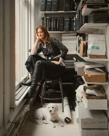 Woman seated on stool next to window in archival shelving room with small white dog lying on the floor below