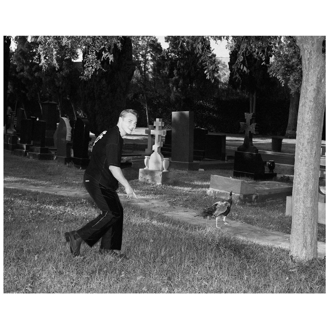 Monochrome shot of a young man gesturing at a peacock in a cemetery with grave markers and trees in the background on a sunny day.