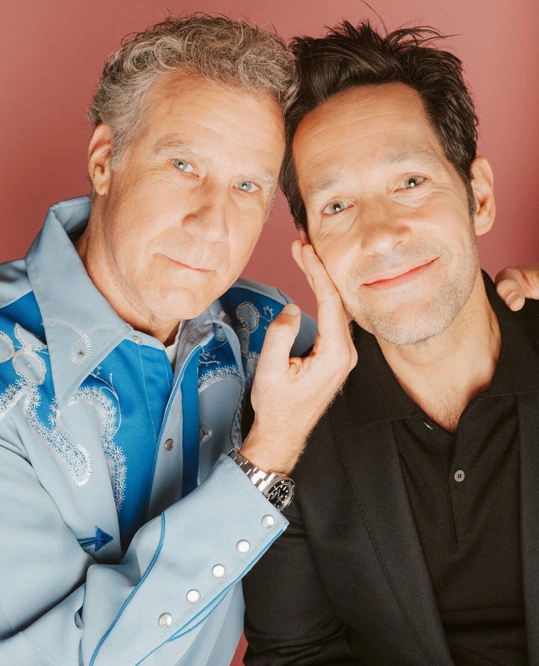 Two adult men pose indoors against a pink background. One is fair-skinned with curly gray hair, wearing an embroidered western shirt; the other, dark hair, black shirt and suit coat, touches his face.