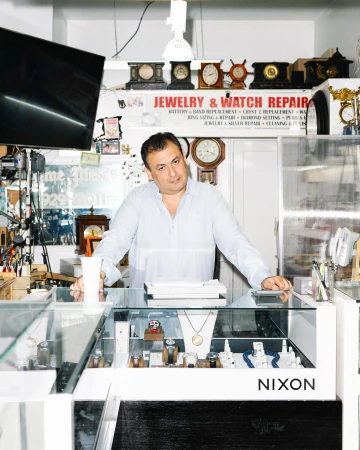 Man in light blue shirt standing behind jewelry and watch repair shop counter, showcasing watches, jewelry, and the Nixon logo.
