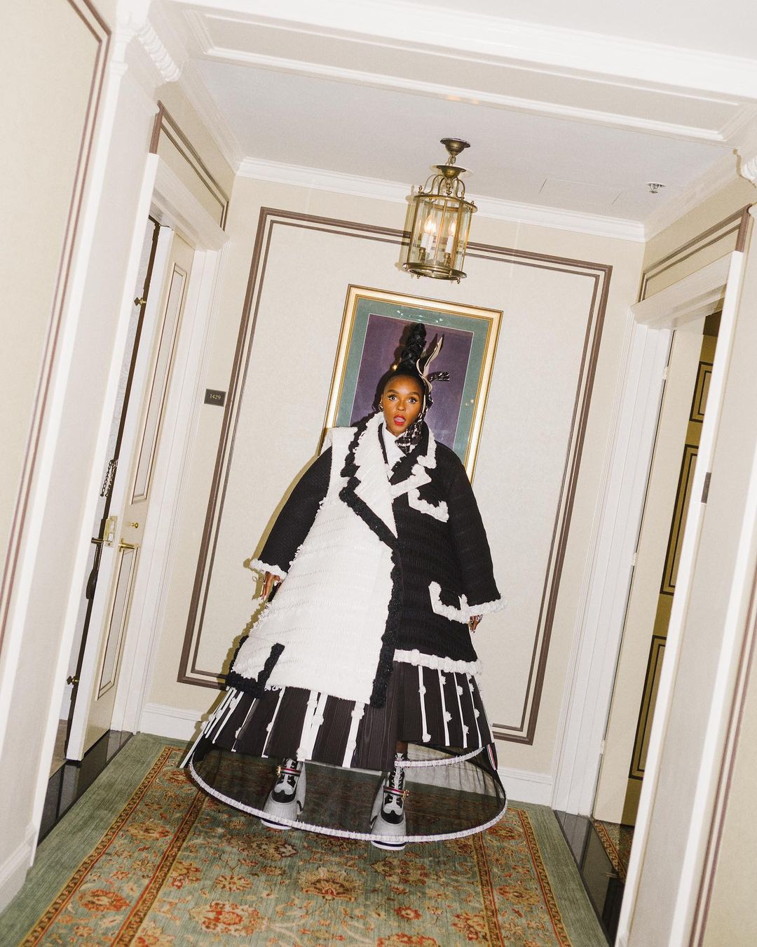Janelle Monáe poses in a black-and-white outfit inside a hallway with ornate details, gold accents, and a vintage rug.