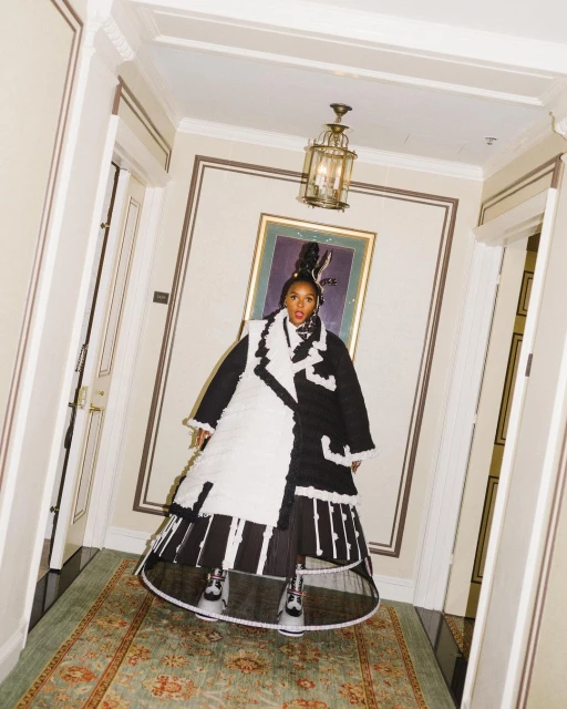 Janelle Monáe poses in a black-and-white outfit inside a hallway with ornate details, gold accents, and a vintage rug.