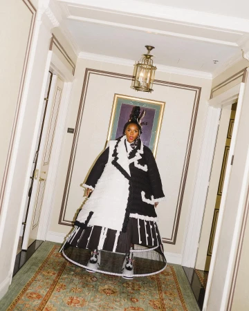 Janelle Monáe poses in a black-and-white outfit inside a hallway with ornate details, gold accents, and a vintage rug.