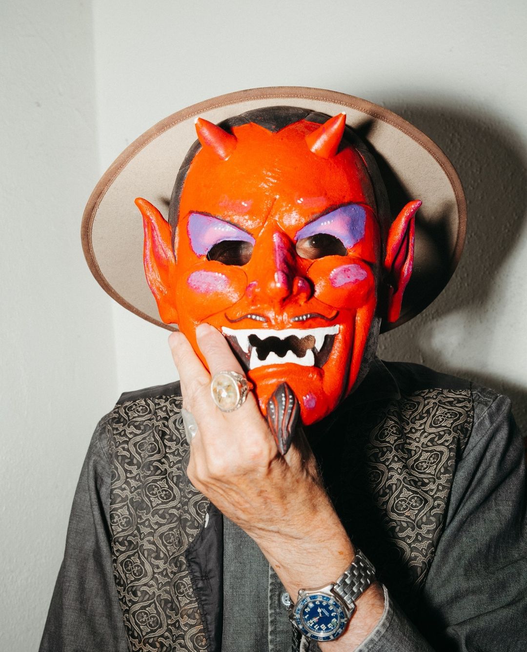 A person wearing a red devil mask and a hat indoors, with visible rings and a watch, creates a unique and theatrical image.