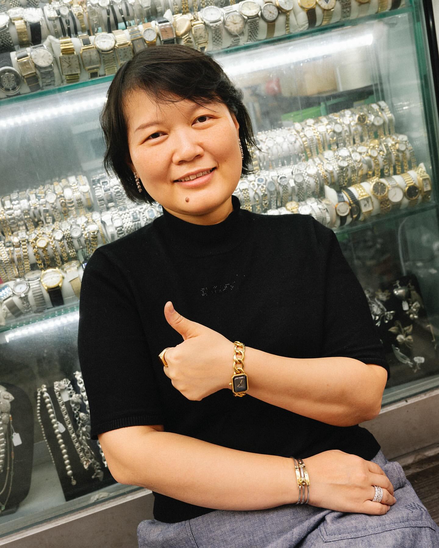 Adult woman smiles, wearing a black top, gold watch and rings, poses in front of a display case full of watches.