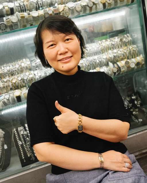 Adult woman smiles, wearing a black top, gold watch and rings, poses in front of a display case full of watches.
