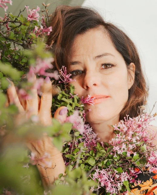 Portrait of a woman with dark hair and floral clothing, surrounded by pink flowers and green leaves in a close-up shot.
