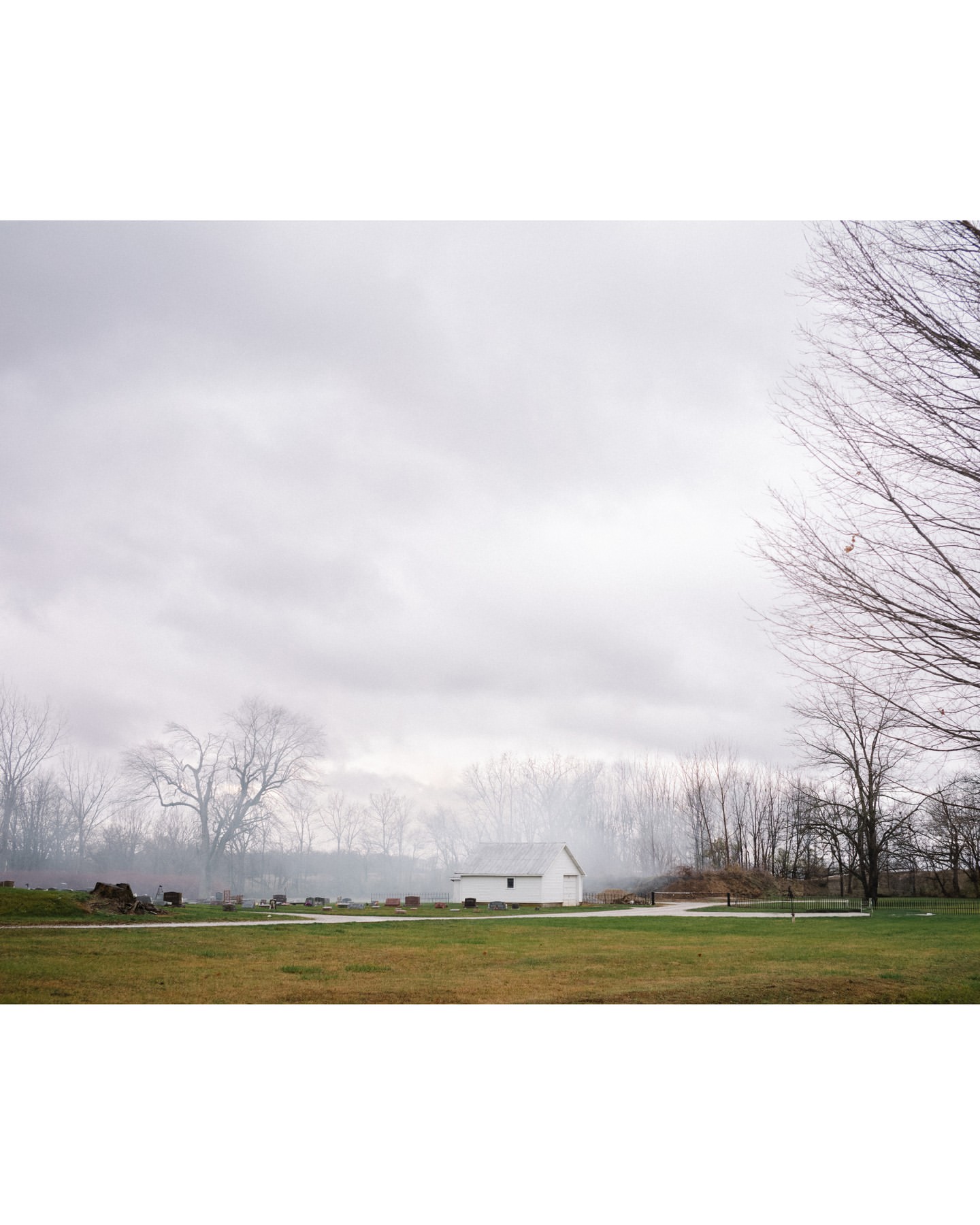 Rural landscape featuring a small white house and a grassy field under a cloudy sky, framed by bare trees in soft, overcast light.