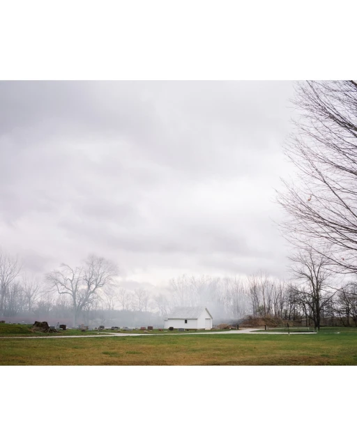 Rural landscape featuring a small white house and a grassy field under a cloudy sky, framed by bare trees in soft, overcast light.