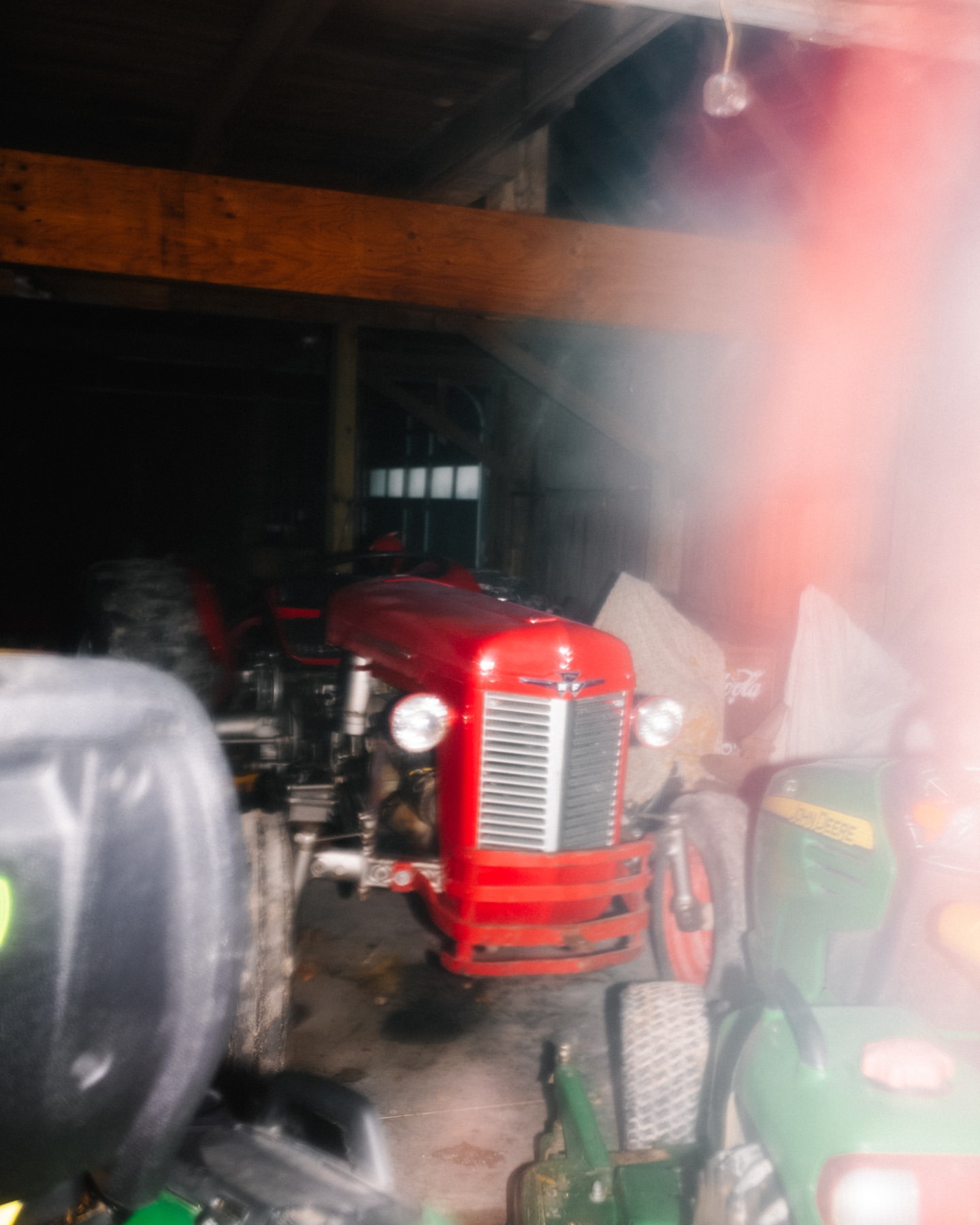 Vintage red tractor parked inside a dimly lit barn with wooden beams and various pieces of farm equipment visible. 