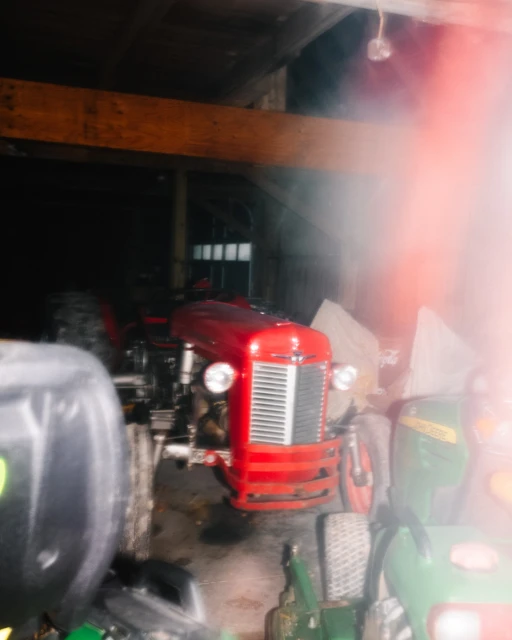 Vintage red tractor parked inside a dimly lit barn with wooden beams and various pieces of farm equipment visible. 