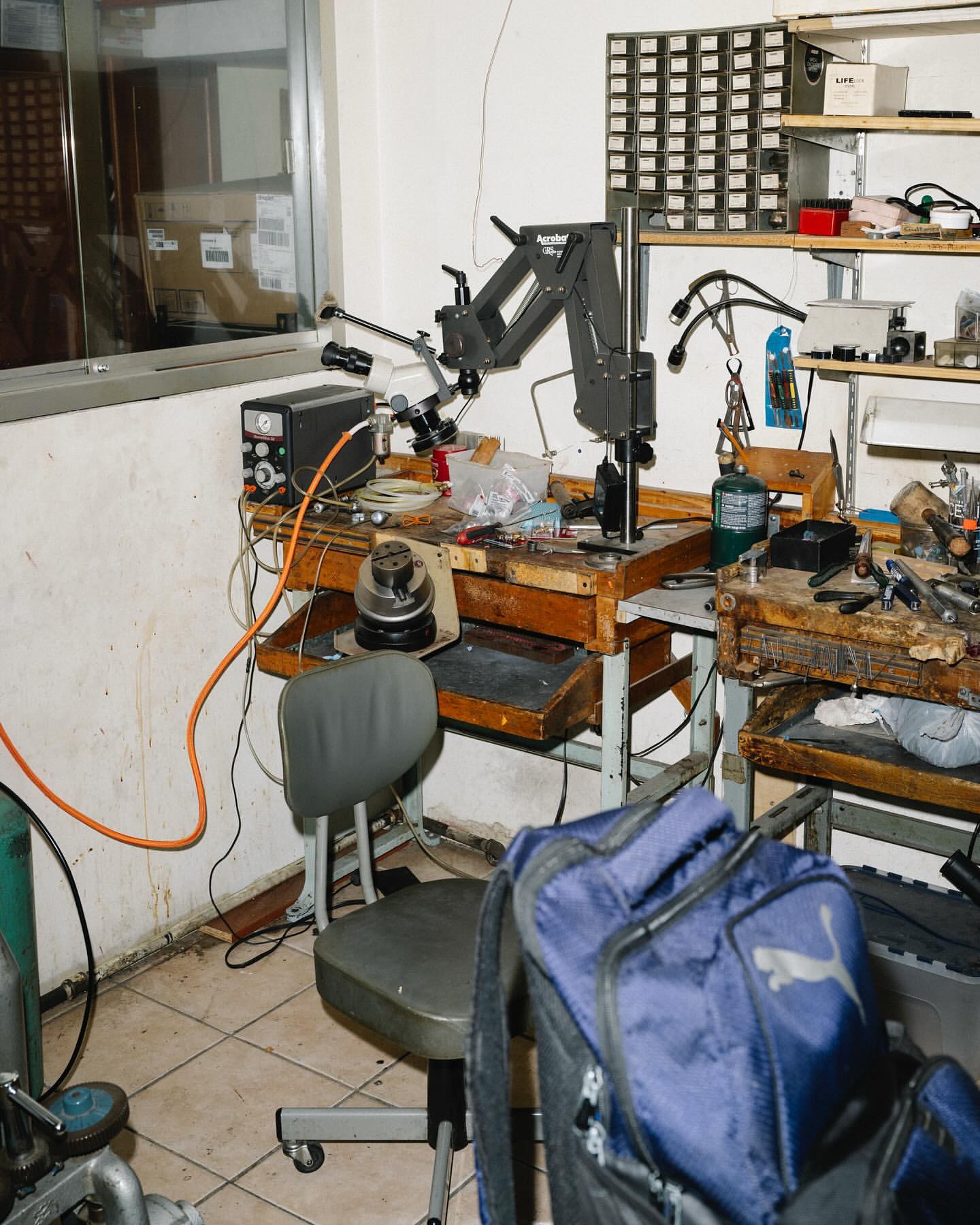 Workbench in a workshop with tools, a microscope, and a blue Puma backpack. The room has a cluttered appearance and industrial vibe.