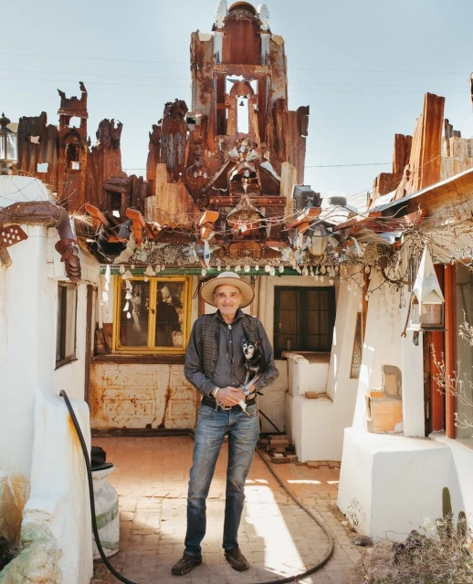 Portrait of an adult man in a straw hat and jeans, holding a small dog outside a unique art installation, under bright sunlight.