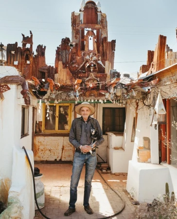 Portrait of an adult man in a straw hat and jeans, holding a small dog outside a unique art installation, under bright sunlight.