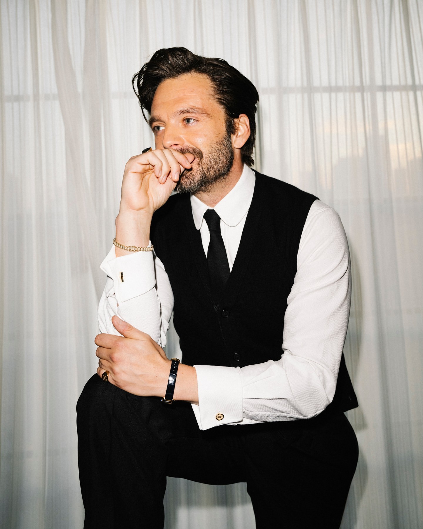 Adult male in black vest and tie poses in an indoor setting with a slight smile and hand near his mouth.
