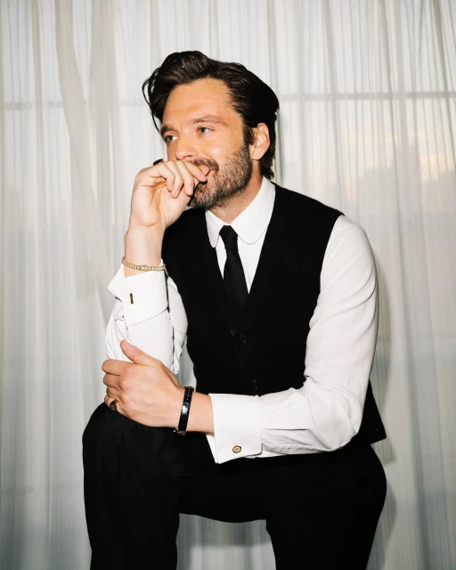 Adult male in black vest and tie poses in an indoor setting with a slight smile and hand near his mouth.