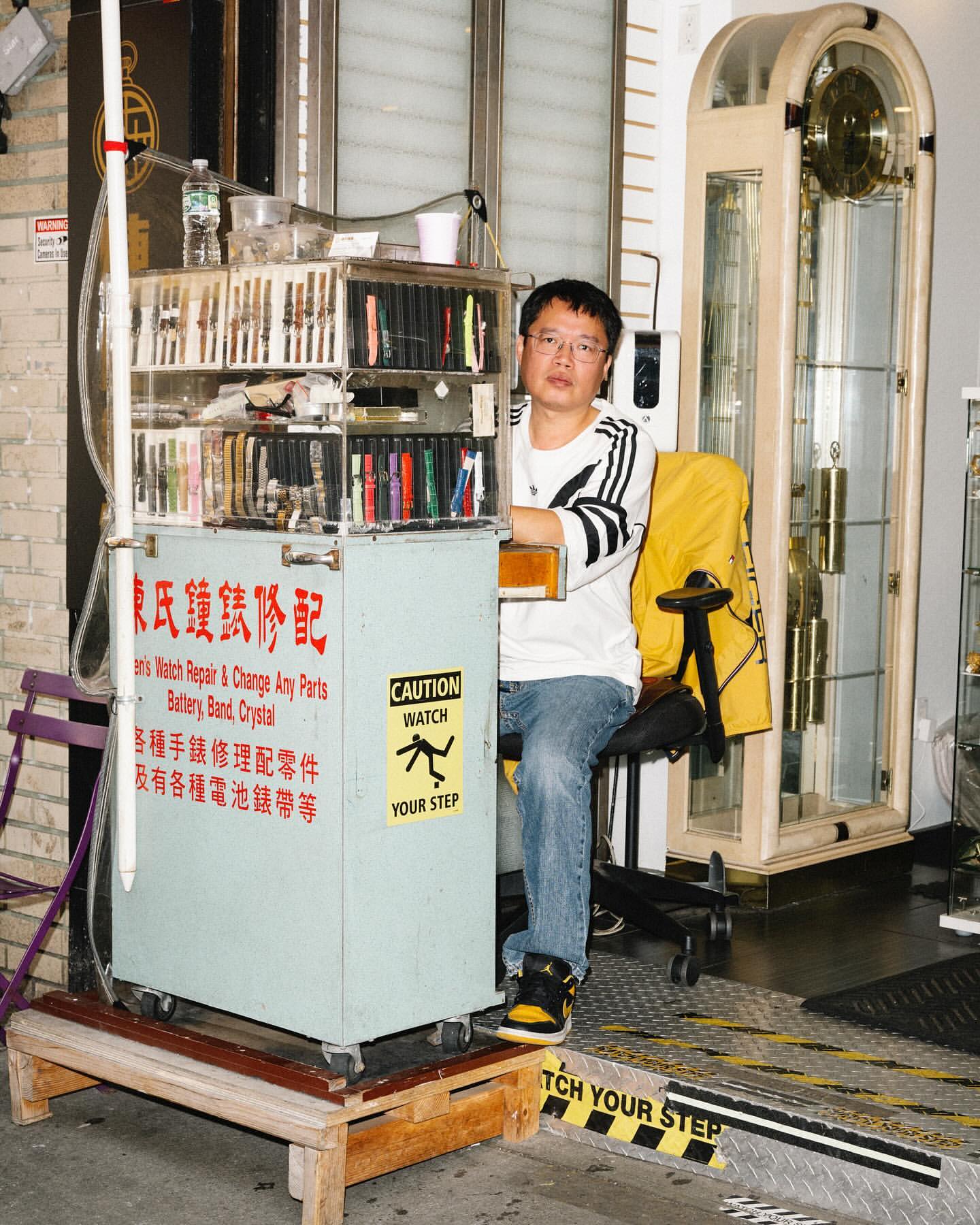Asian male watch repairer sitting at his mobile workbench displaying a variety of watch straps and parts, inside what appears to be a jewelry store.