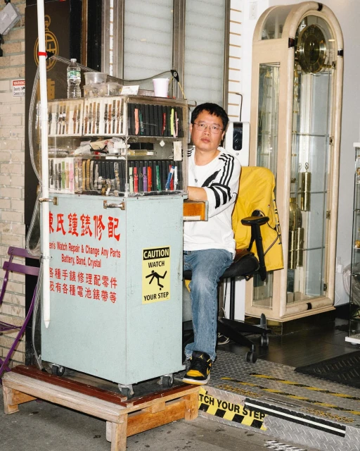 Asian male watch repairer sitting at his mobile workbench displaying a variety of watch straps and parts, inside what appears to be a jewelry store.
