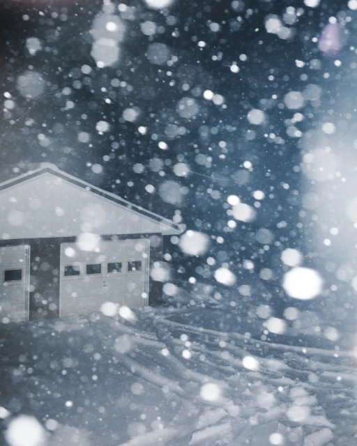 Snowy landscape featuring a building with closed garage doors, with falling snow creating bokeh effects against a dark background.