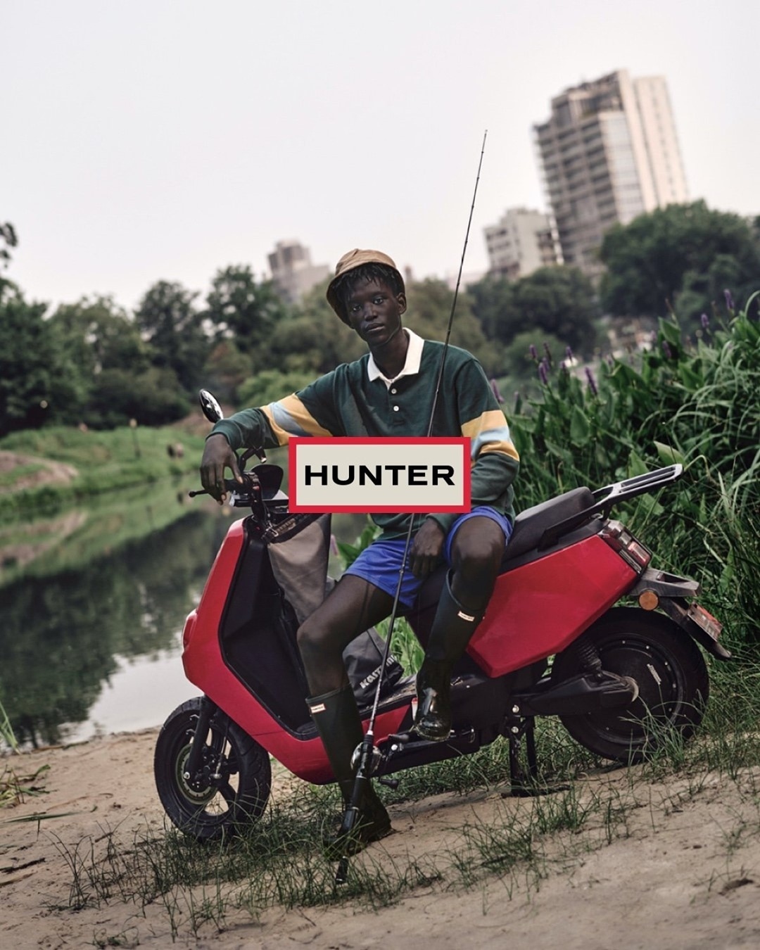 A person in a hat and green shirt sitting on a red scooter next to a body of water with trees and city buildings in the background.