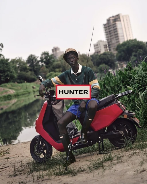 A person in a hat and green shirt sitting on a red scooter next to a body of water with trees and city buildings in the background.