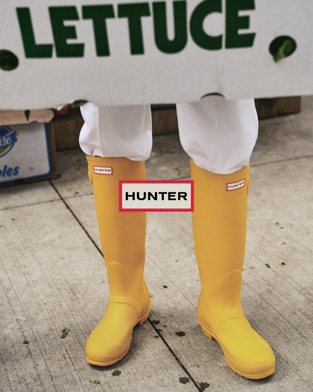 Person wears yellow Hunter boots while carrying a box of lettuce. The shot is at eye-level outside on the sidewalk.