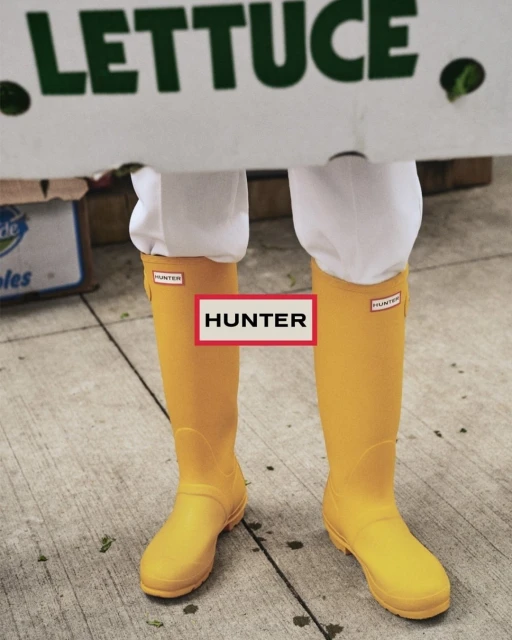 Person wears yellow Hunter boots while carrying a box of lettuce. The shot is at eye-level outside on the sidewalk.