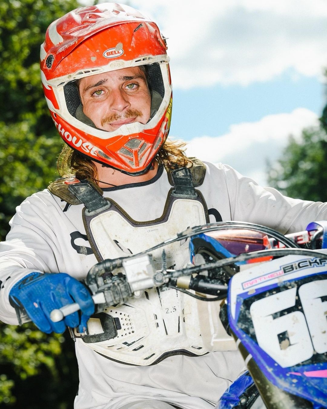 Male motorcyclist wearing helmet and chest protector pauses on a dirt bike in an outdoor setting with green foliage and partly cloudy skies.
