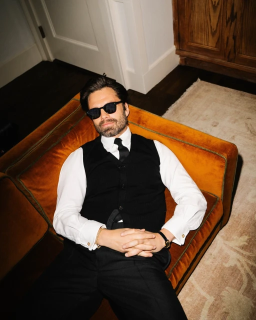 Man in a black suit and sunglasses poses on an orange velvet couch in an indoor setting with neutral walls and flooring.