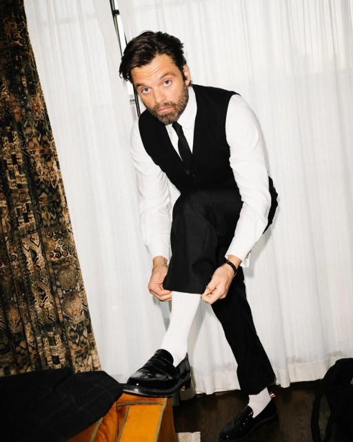A man in a black vest and pants sits on a chair, pulling up his white socks and black loafers in a bright, indoor setting.