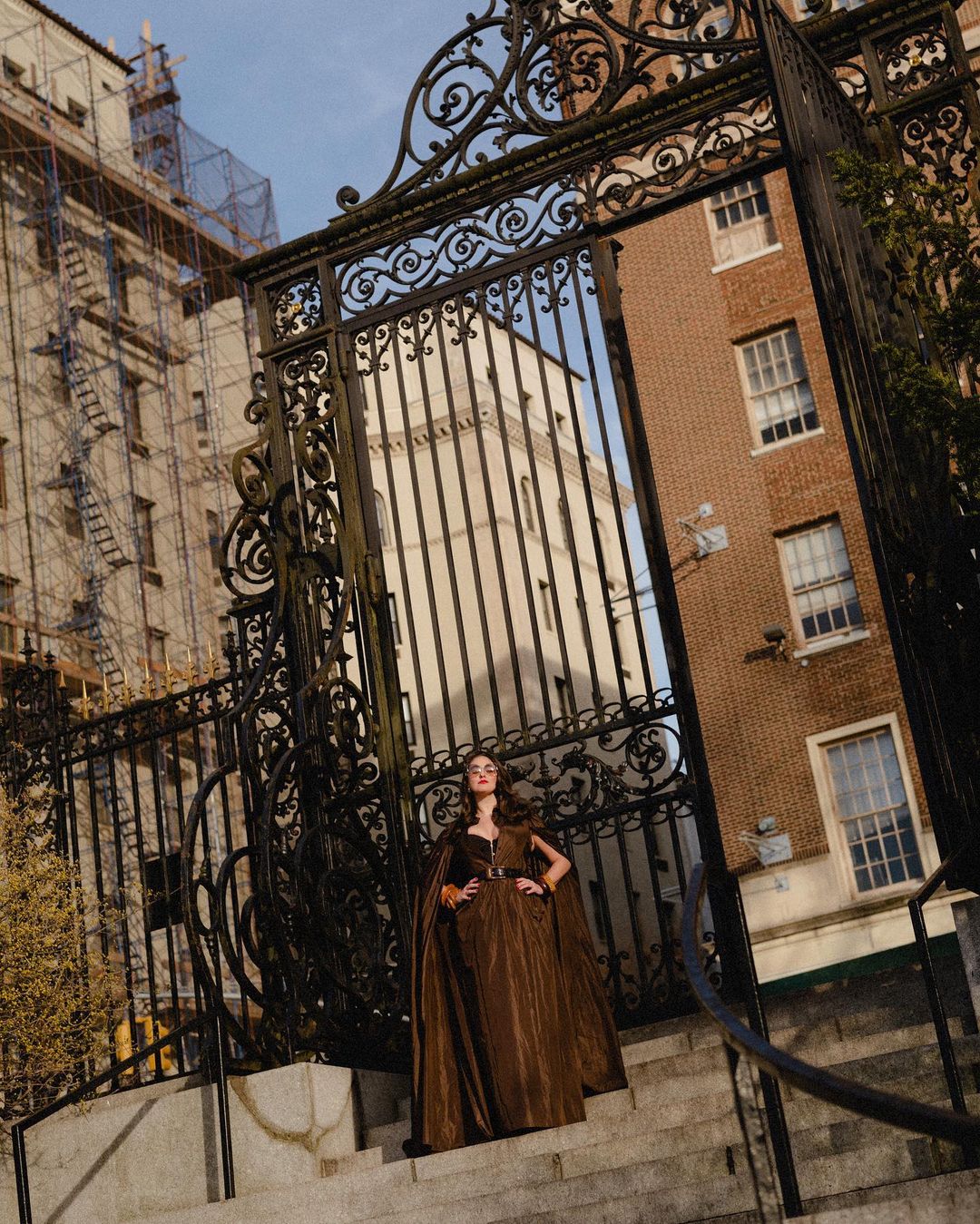 A woman in a brown dress stands on steps behind a wrought iron gate, with brick buildings in the background on a sunny day.