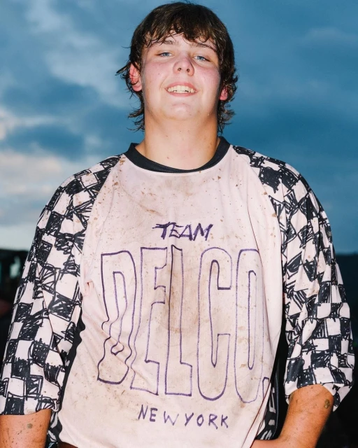 A young person with short brown hair smiles in a close-up shot, wearing a white jersey with a black and white checkered sleeve pattern.