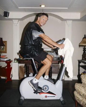 An adult male is riding an exercise bike indoors wearing a black plastic bag as a makeshift sweat suit, with living room decor in the background.