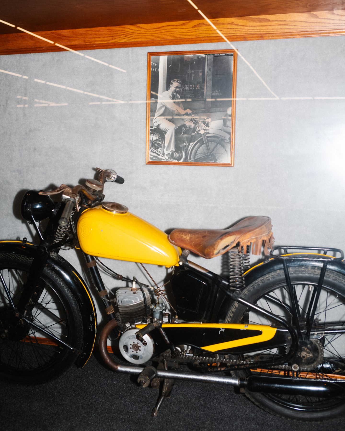 Vintage motorcycle with yellow tank and brown seat, displayed indoors with a framed black and white photo in the background.