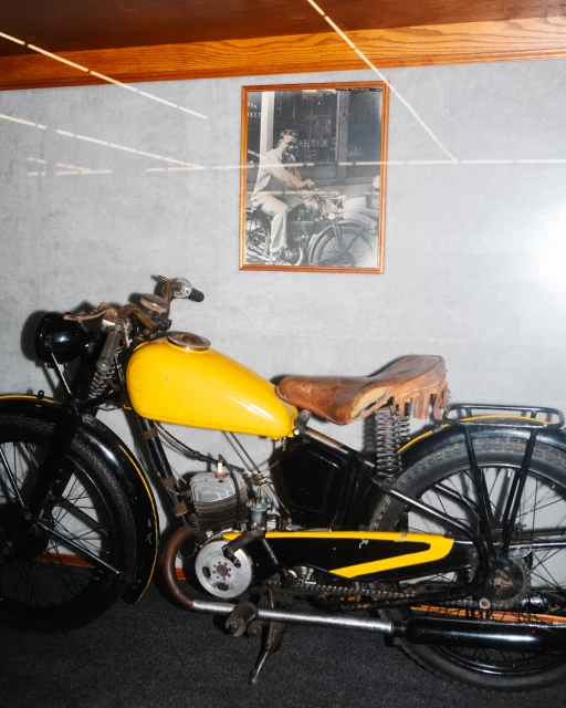 Vintage motorcycle with yellow tank and brown seat, displayed indoors with a framed black and white photo in the background.
