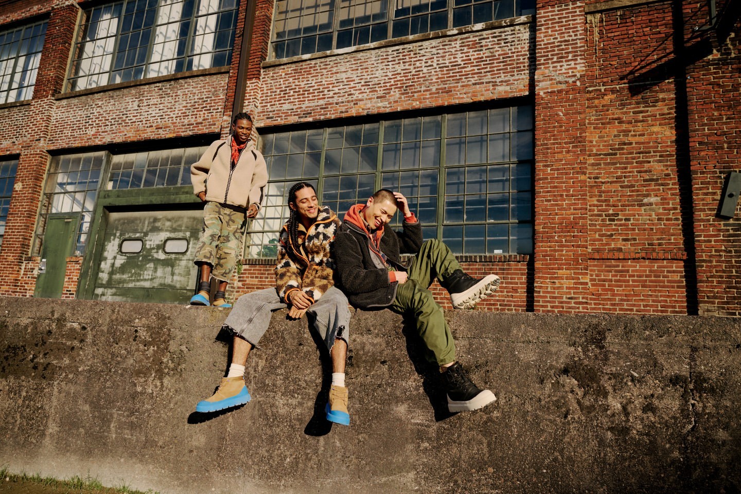 Three people in outerwear are sitting and standing on a concrete ledge in front of a brick building with large windows and a garage door.