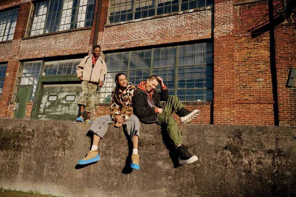 Three people in outerwear are sitting and standing on a concrete ledge in front of a brick building with large windows and a garage door.