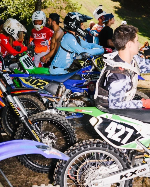 Motocross riders on dirt bikes pause with helmets on near a wooded area, captured at eye-level with clear focus on the bikes and riders.