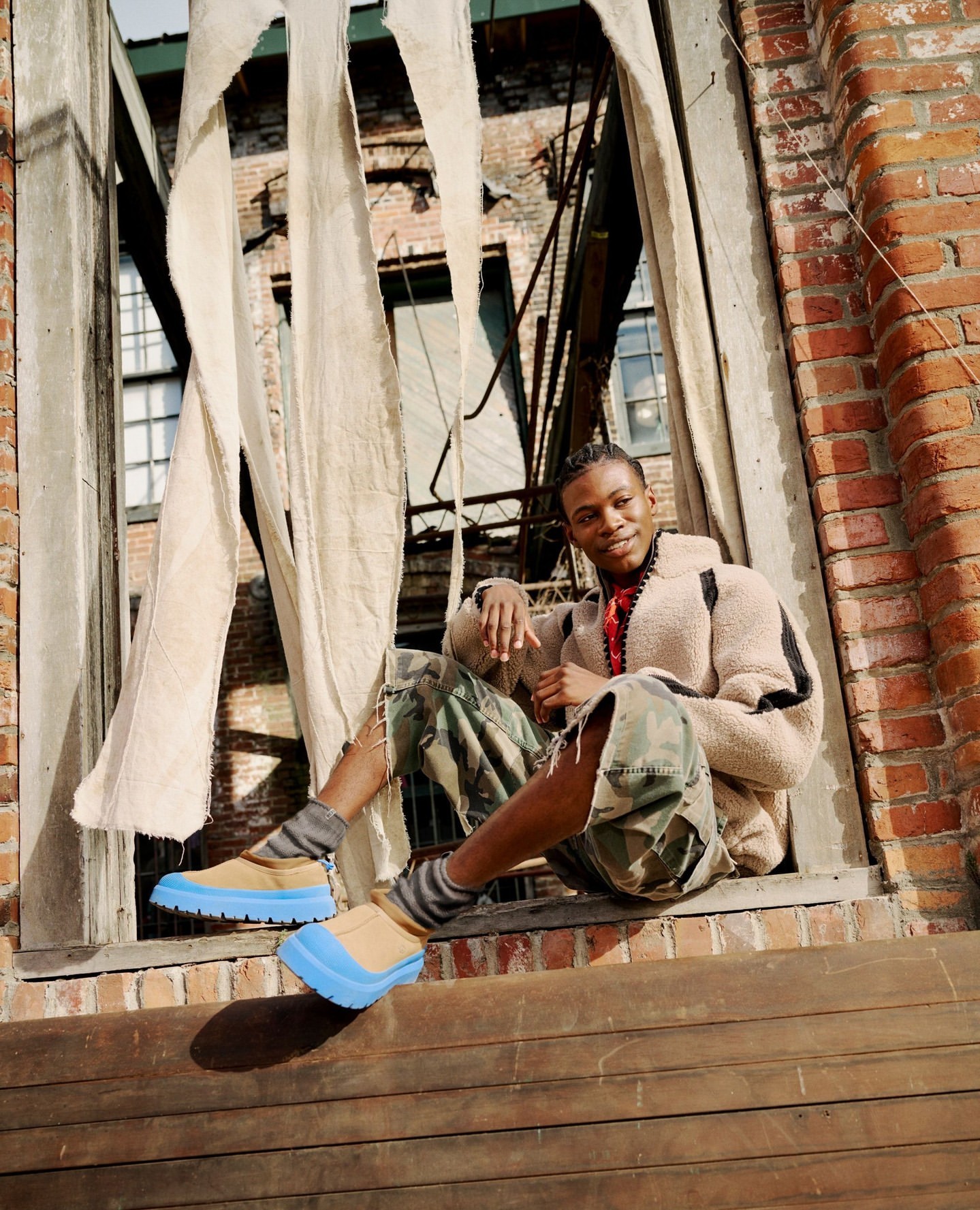 A person sits on a window ledge in a brown jacket and patterned shorts, with a brick building and draped fabric in the background.