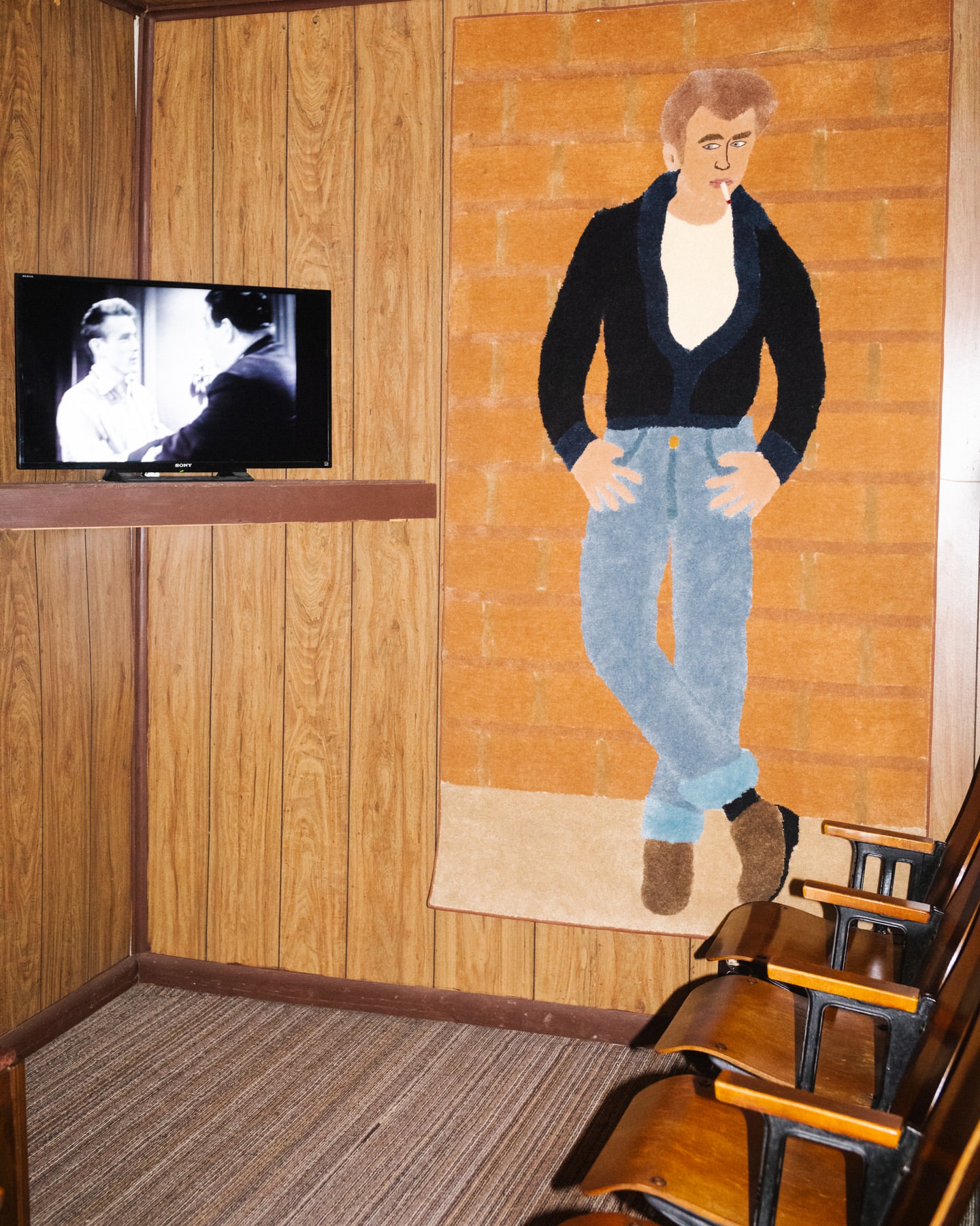Hand-crafted art piece depicting James Dean in jeans and black jacket smoking a cigarette, hanging in a recreation room with theatre chairs.