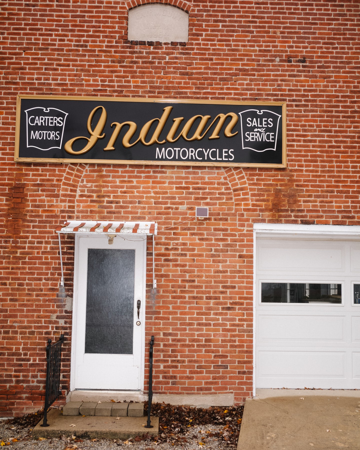 Exterior of a brick building featuring an Indian Motorcycles sign, a white door with a small awning, and a white garage door.