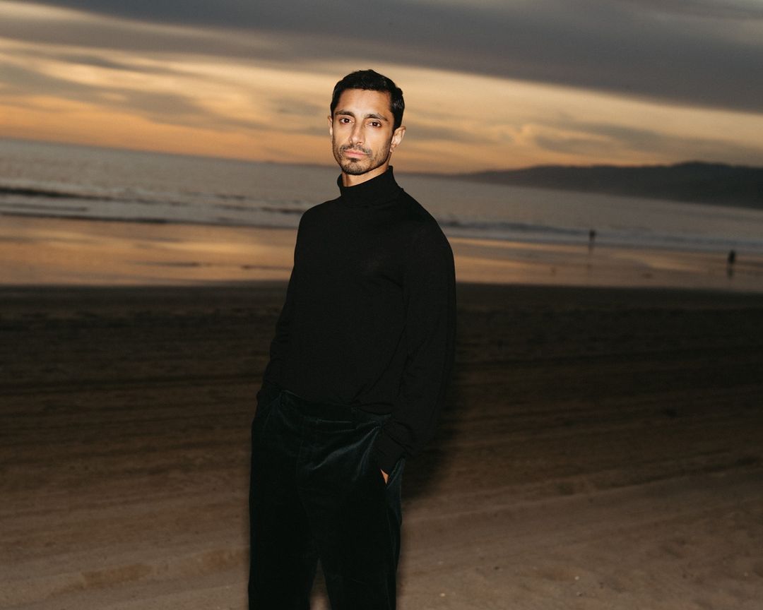 An adult male stands on a sandy beach at sunset, wearing a black turtleneck sweater and dark trousers, with ocean waves behind him.