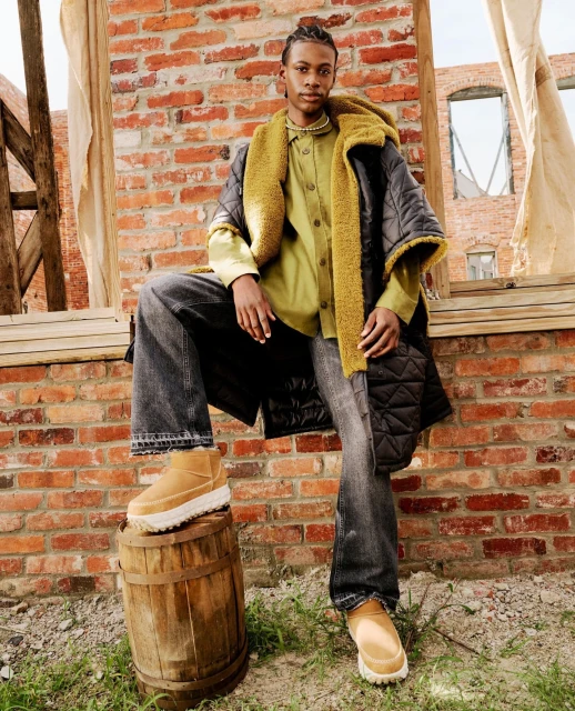A stylish individual poses confidently outdoors against a rustic brick backdrop, showcasing fashion-forward clothing and footwear.