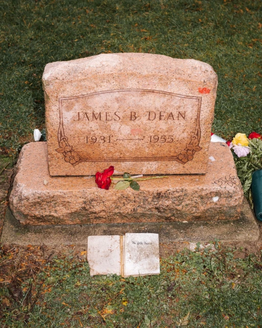 James Dean's grave marker with a rose and an open book with text lies on the ground in front of the tombstone on a grassy lawn.