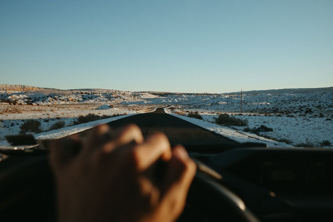 Point-of-view shot of a driver's hands on the steering wheel, looking out over a long road through a snowy desert landscape under a blue sky.