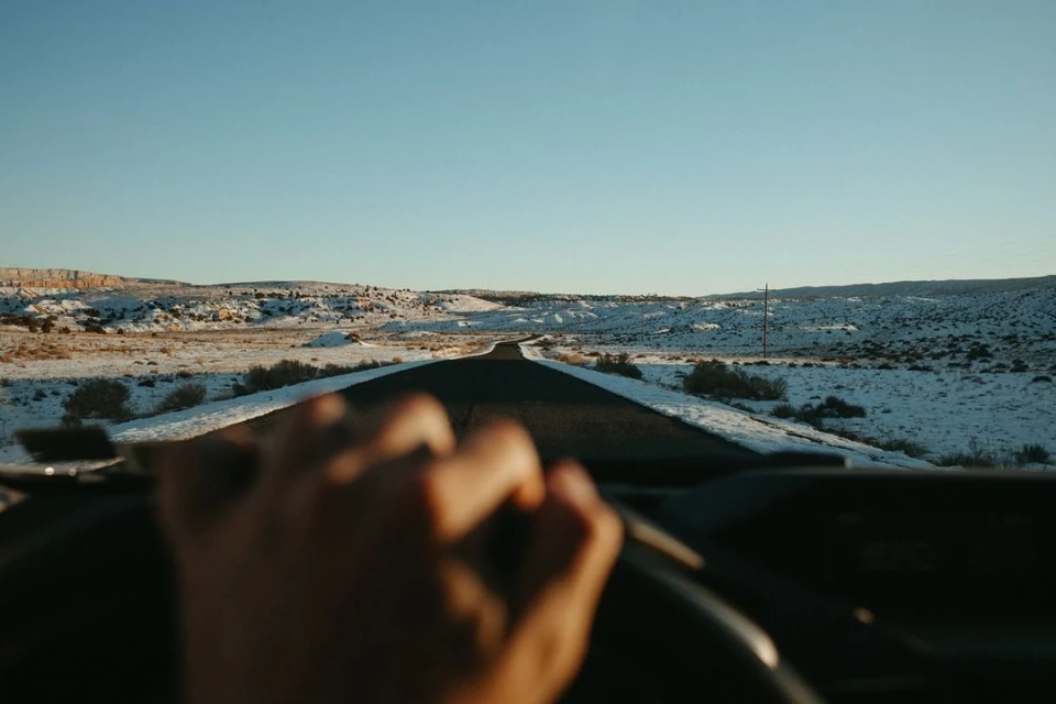 Point-of-view shot of a driver's hands on the steering wheel, looking out over a long road through a snowy desert landscape under a blue sky.