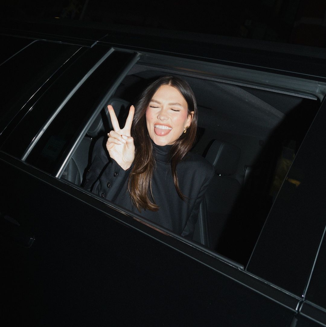 A young adult woman with long brown hair leans out the window of a black car making a peace sign with her fingers. She wears black clothing.