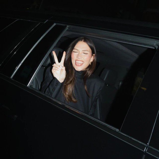 A young adult woman with long brown hair leans out the window of a black car making a peace sign with her fingers. She wears black clothing.