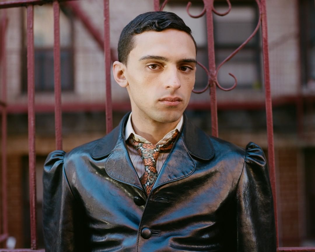 Portrait of a young adult male with short, dark hair wearing a black leather jacket in front of a wrought iron gate outdoors.